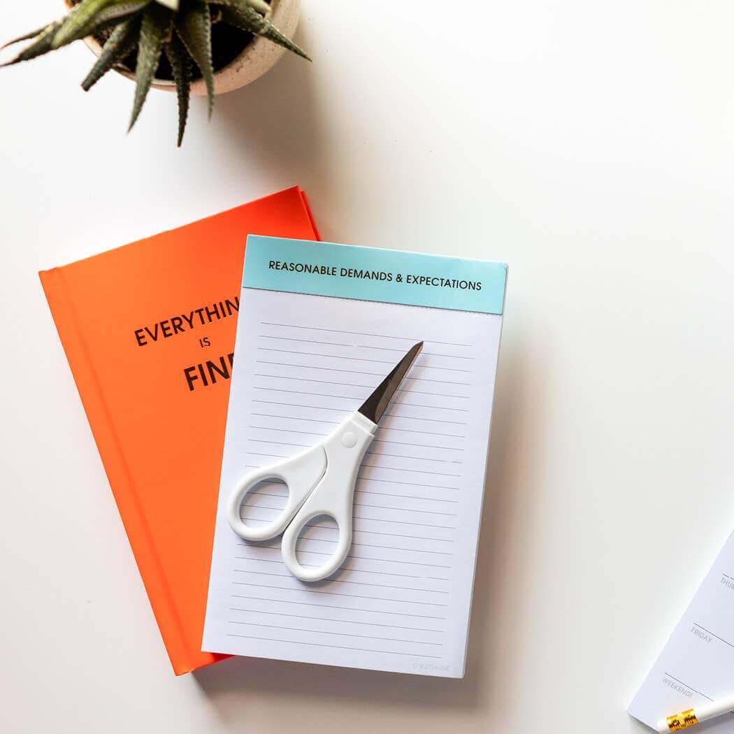 Neon orange "Everything is Fine" journal with gold foil print, lined notepad, white scissors, and a plant on a white desk. Journaling supplies.