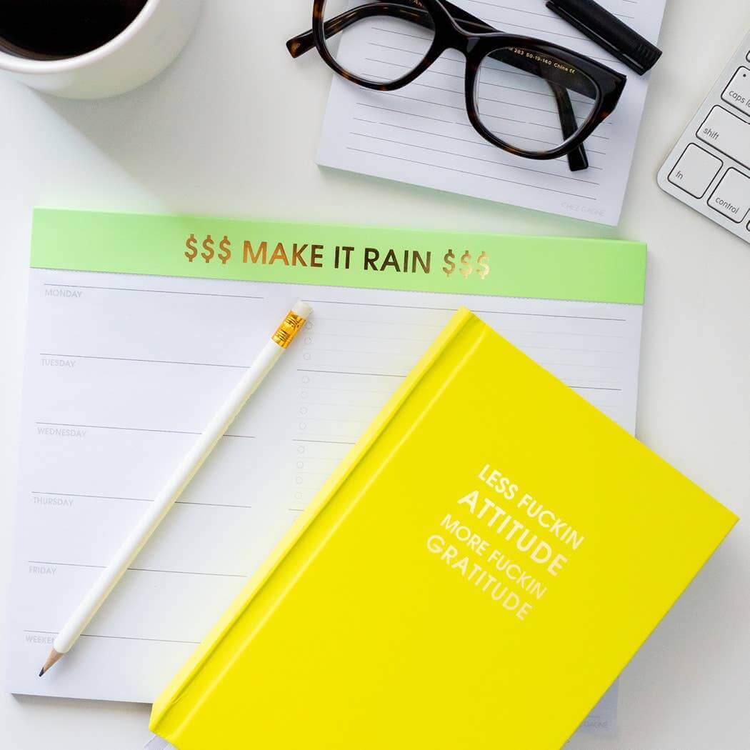 Neon yellow hardcover journal with "Less Attitude More Gratitude" design, on a desk with glasses, coffee, keyboard, and "Make It Rain" notepad.