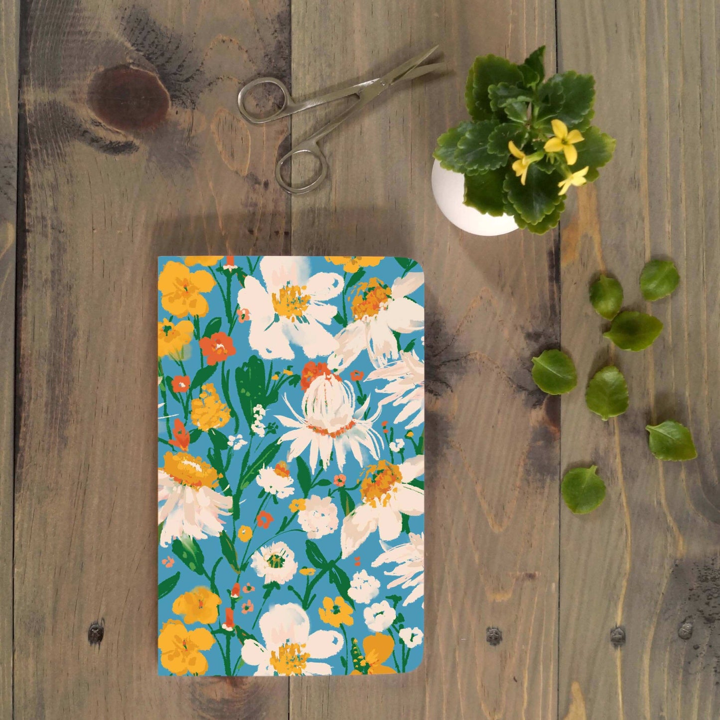 Blue Bouquet Layflat Journal Notebook with floral cover on rustic wooden table, surrounded by green leaves, scissors, and potted plant. Perfect for journaling.