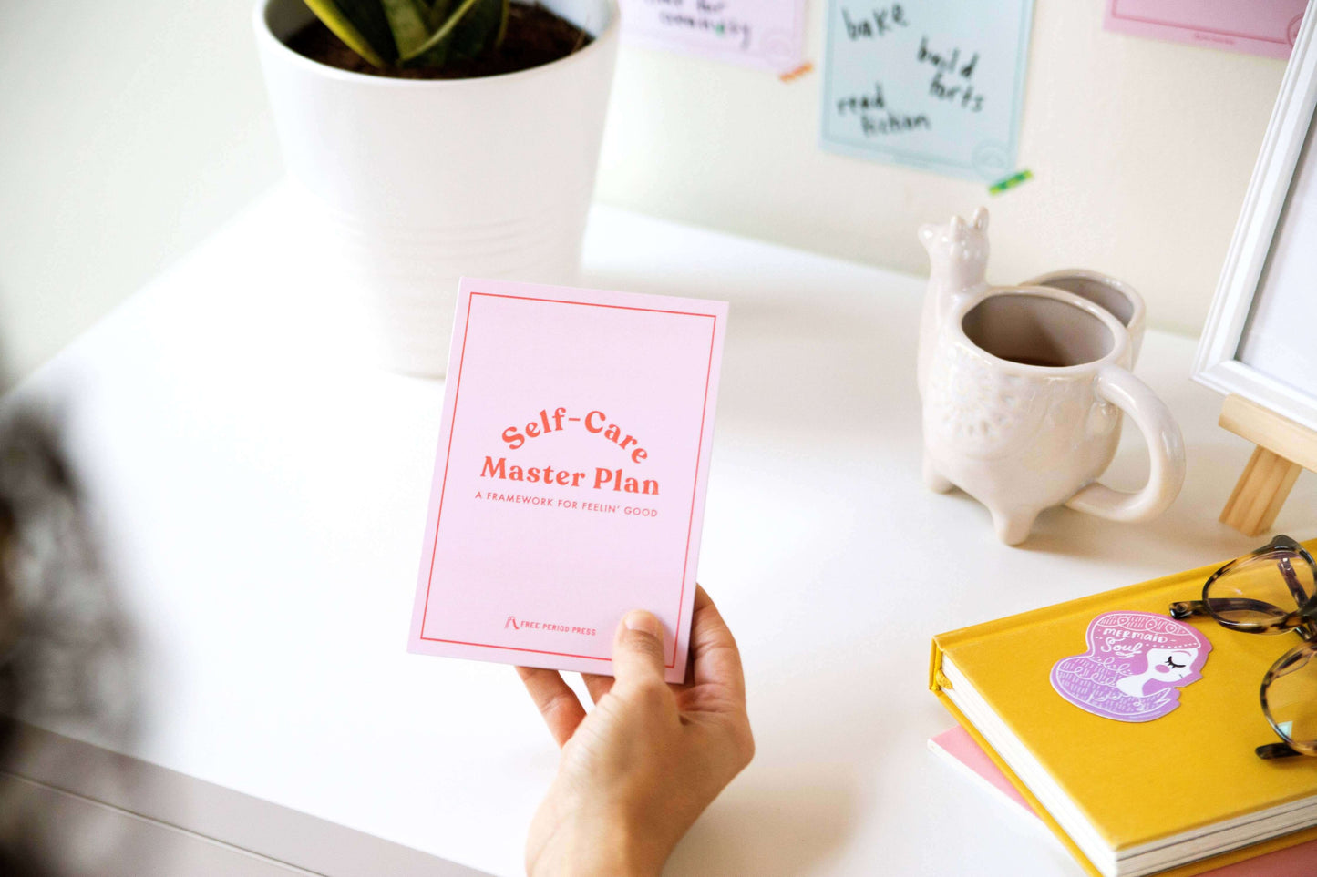 Holding Self-Care Master Plan book for guided journaling with journal supplies on desk