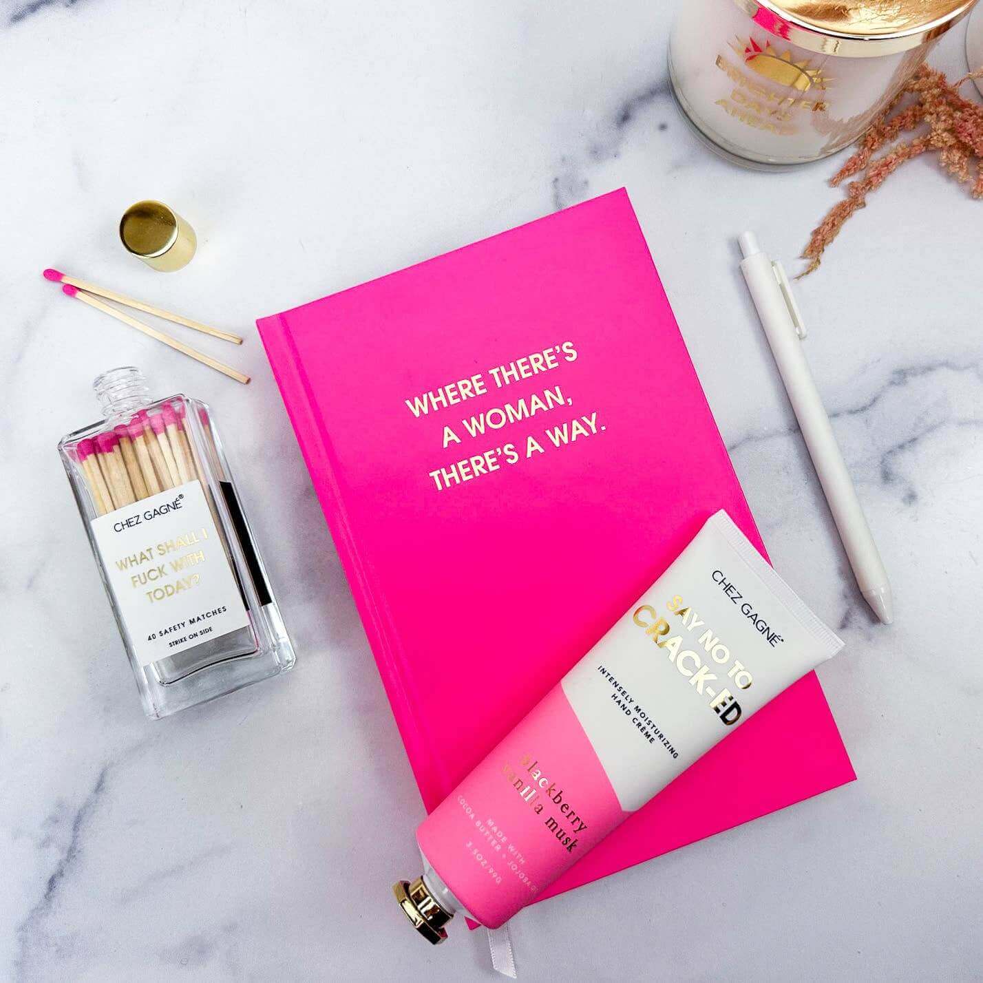 Bright pink 'Where There's A Woman There's A Way' journal with gold foil print, surrounded by matches, lotion, and a pen on a marble surface