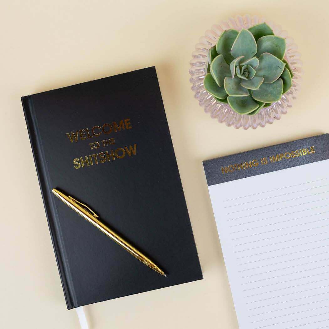 "Welcome to the S***show" bright journal with gold foil print, black hardcover, pen, lined notepad, and succulent for stylish journaling.