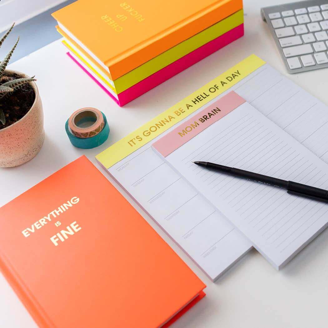 Bright neon orange "Everything is Fine" journal with gold foil print, surrounded by colorful journals, desk supplies, and note pads.