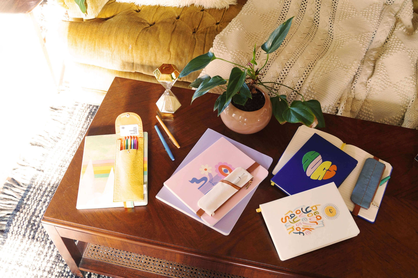 Collection of colorful notebooks and journaling essentials arranged on a wooden table with a leafy plant and cozy yellow couch in the background.