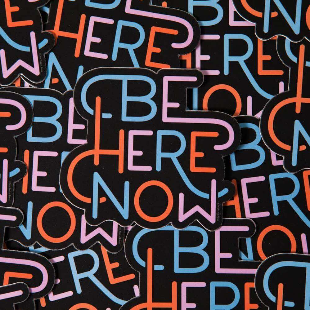 Bright and colorful "Be Here Now" vinyl decal stickers piled up, perfect for laptops, journals, and more - stay present with style!