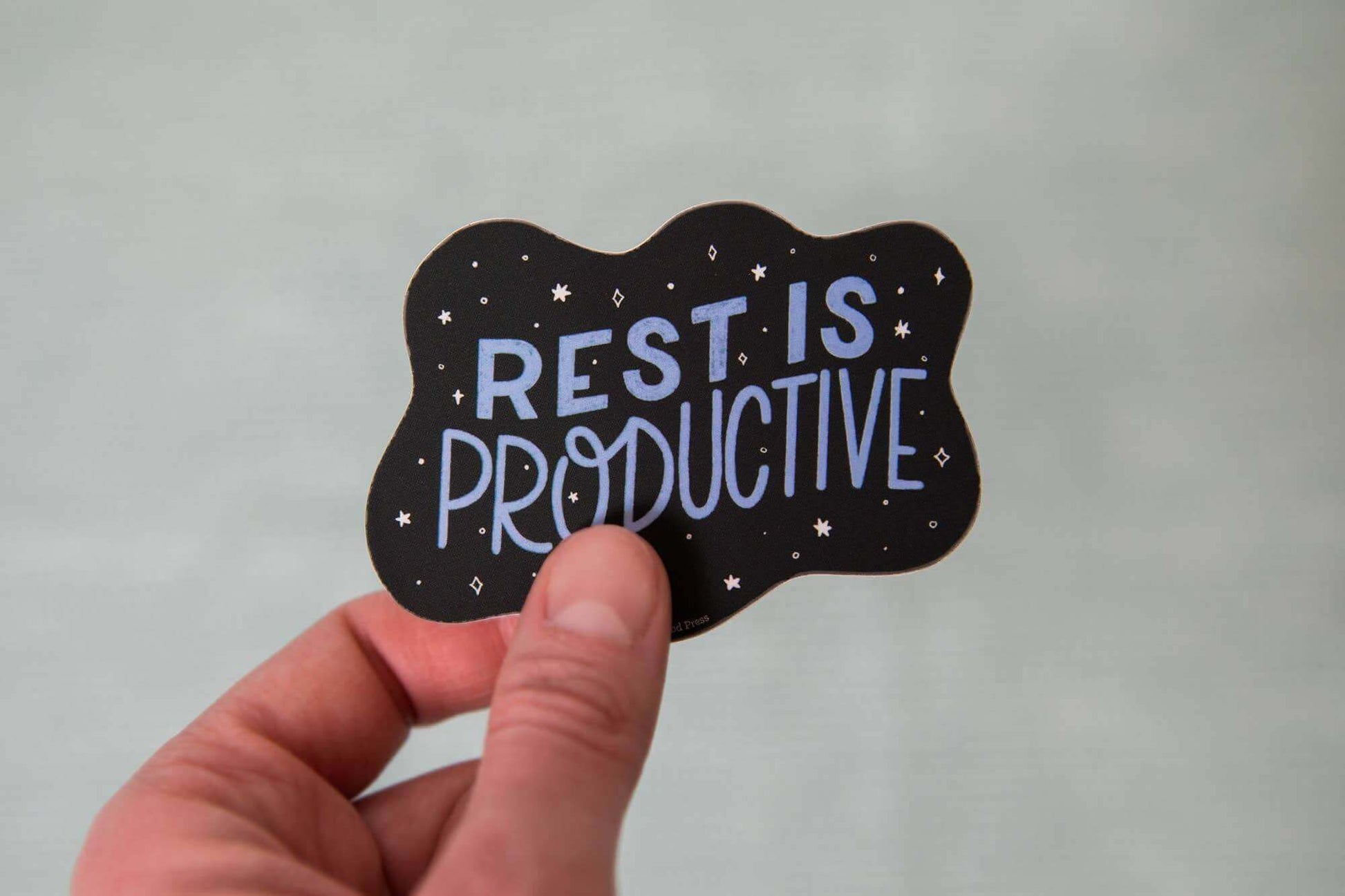 Hand holding a vinyl decal sticker that reads 'Rest is Productive' in bold letters, perfect for laptops, water bottles, and journaling supplies.