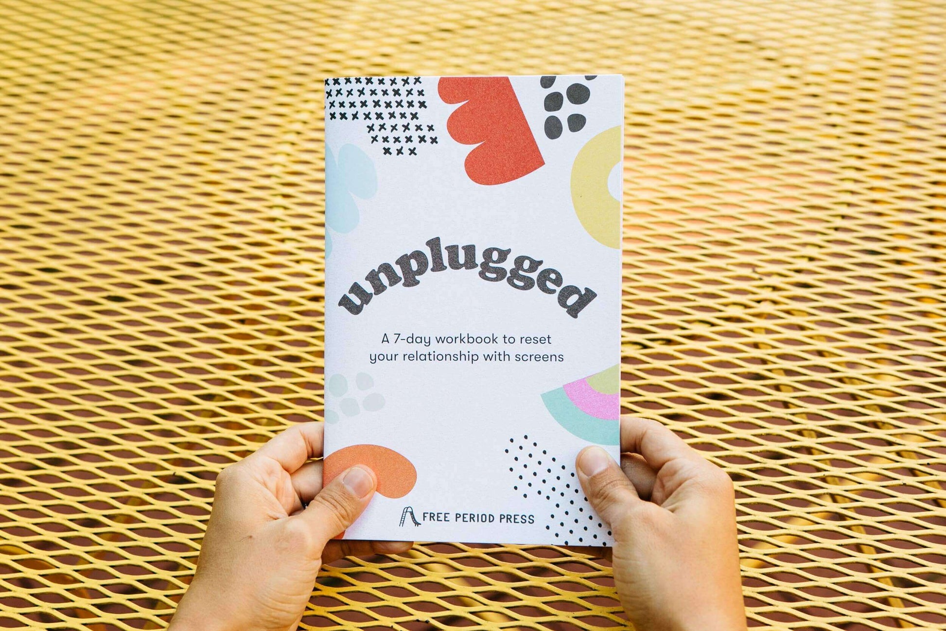 Hands holding 'Unplugged: A 7-day Workbook to Reset Your Relationship with Screens' against a yellow mesh background.