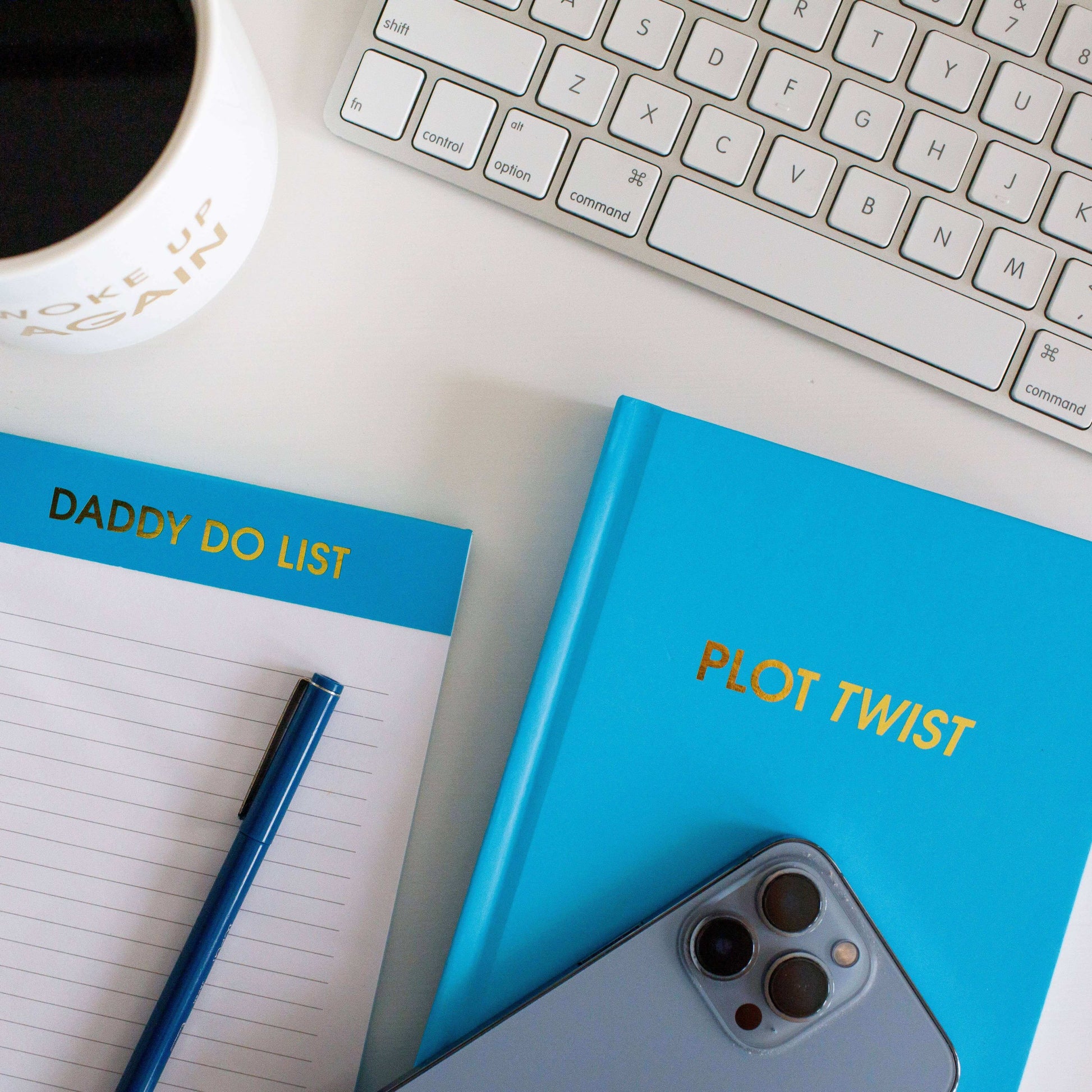 Plot Twist Bright Journal with Blue Cover and Gold Foil Printing next to an iPhone, notebook, and a cup on a white desk for journaling and prompts.