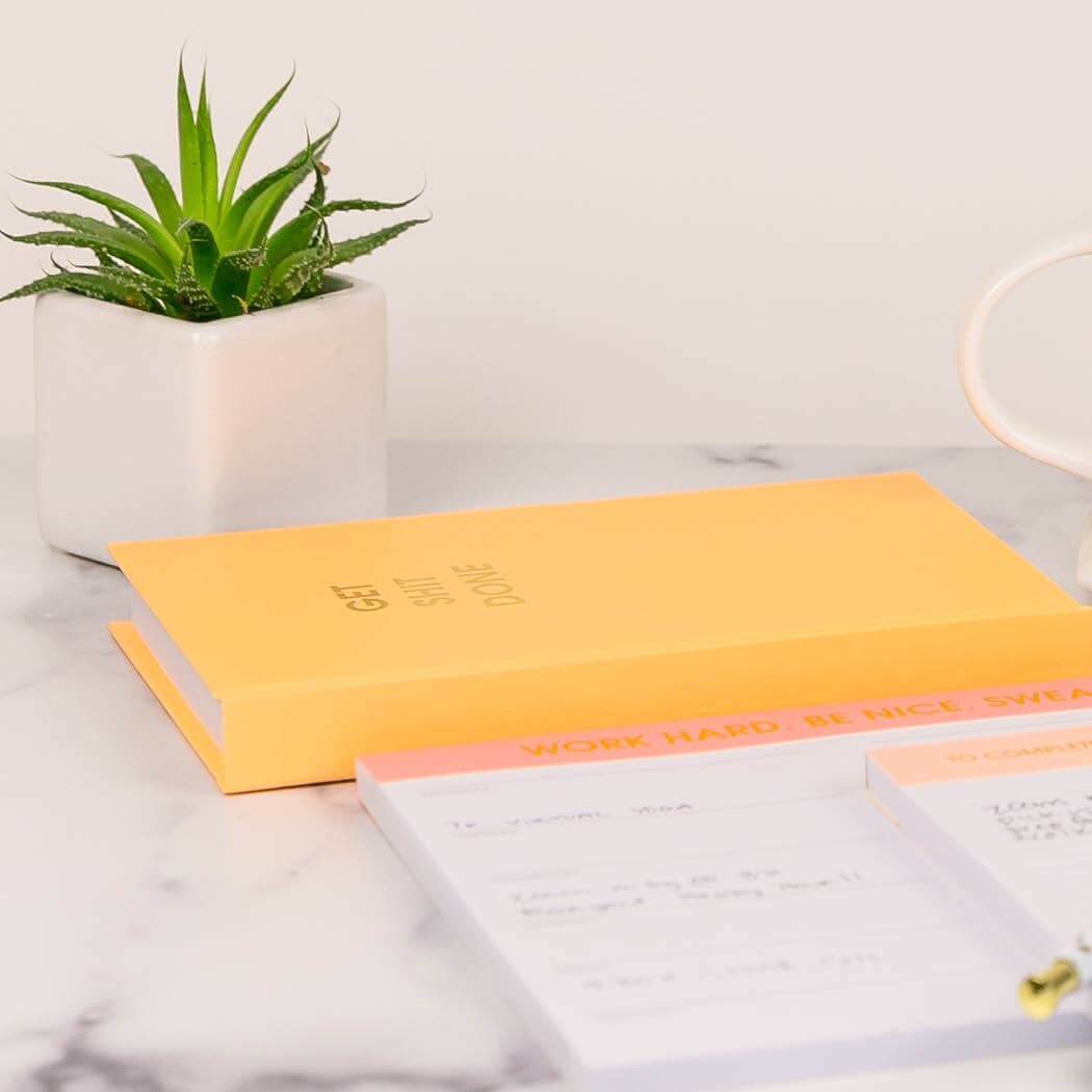 Bright Mimosa Orange hardcover "Get S*** Done" journal with gold foil print, perfect for journaling. Motivational notebook on marble desk.