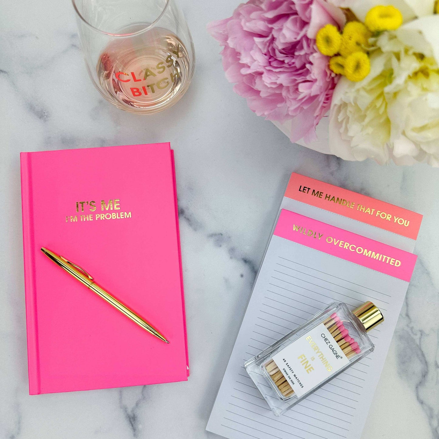 Cosmo Pink "It's Me. I'm The Problem" journal with gold foil, surrounded by a gold pen, pink flowers, notepads, and a glass on marble.