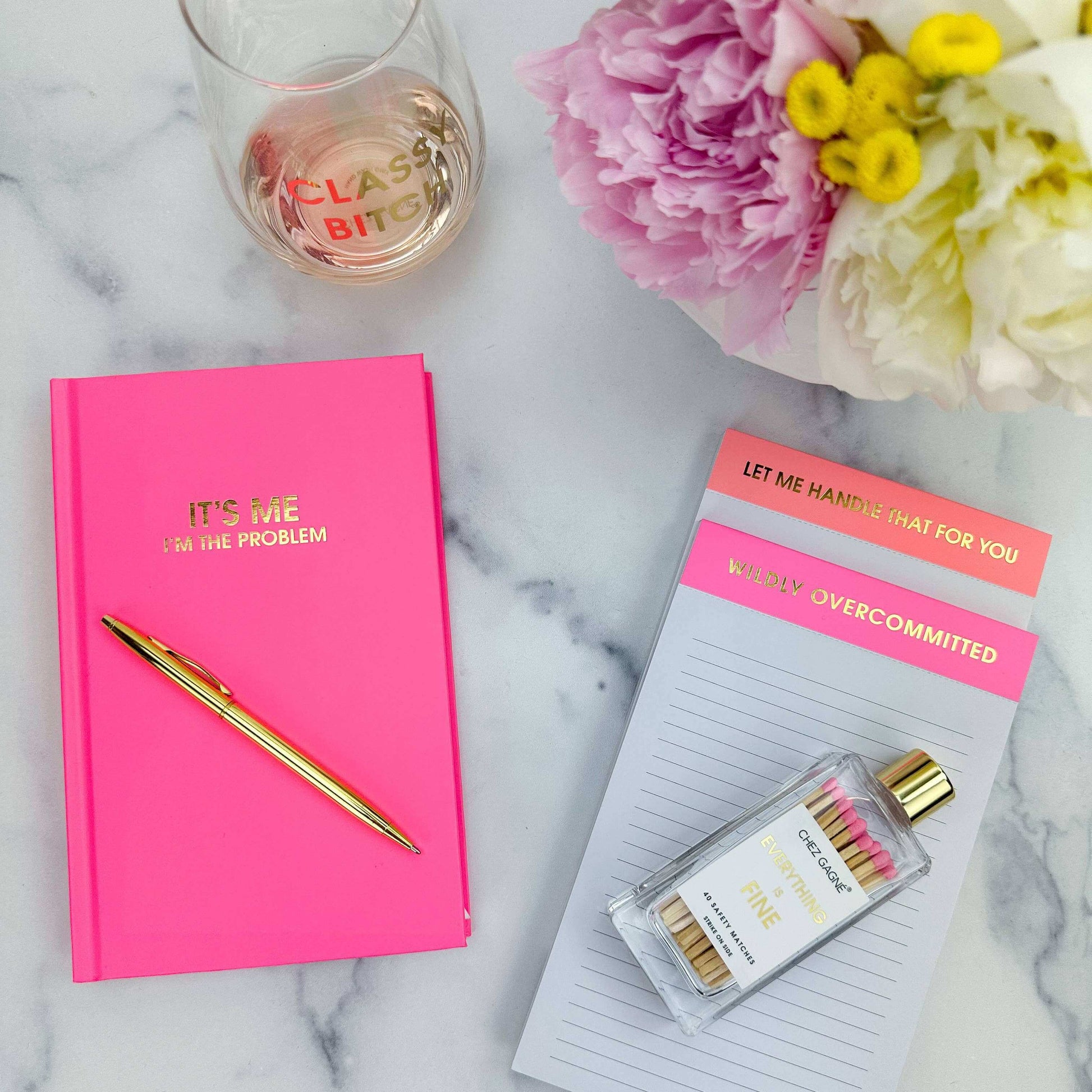 Cosmo Pink "It's Me. I'm The Problem" journal with gold foil, surrounded by a gold pen, pink flowers, notepads, and a glass on marble.