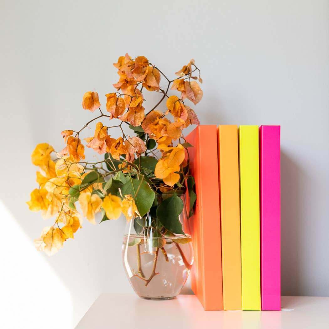 Bright neon journals in orange, yellow, and pink standing next to a vase of yellow flowers on a white shelf, perfect for journaling.