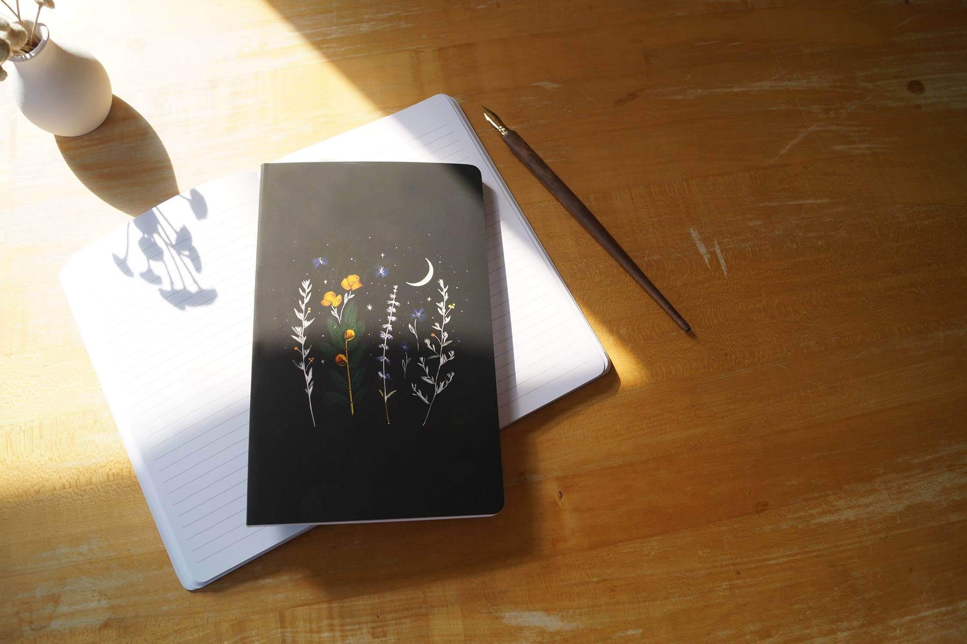 Black notebook with floral design on open notebook on wooden desk with pen and shadow of vase in sunlight