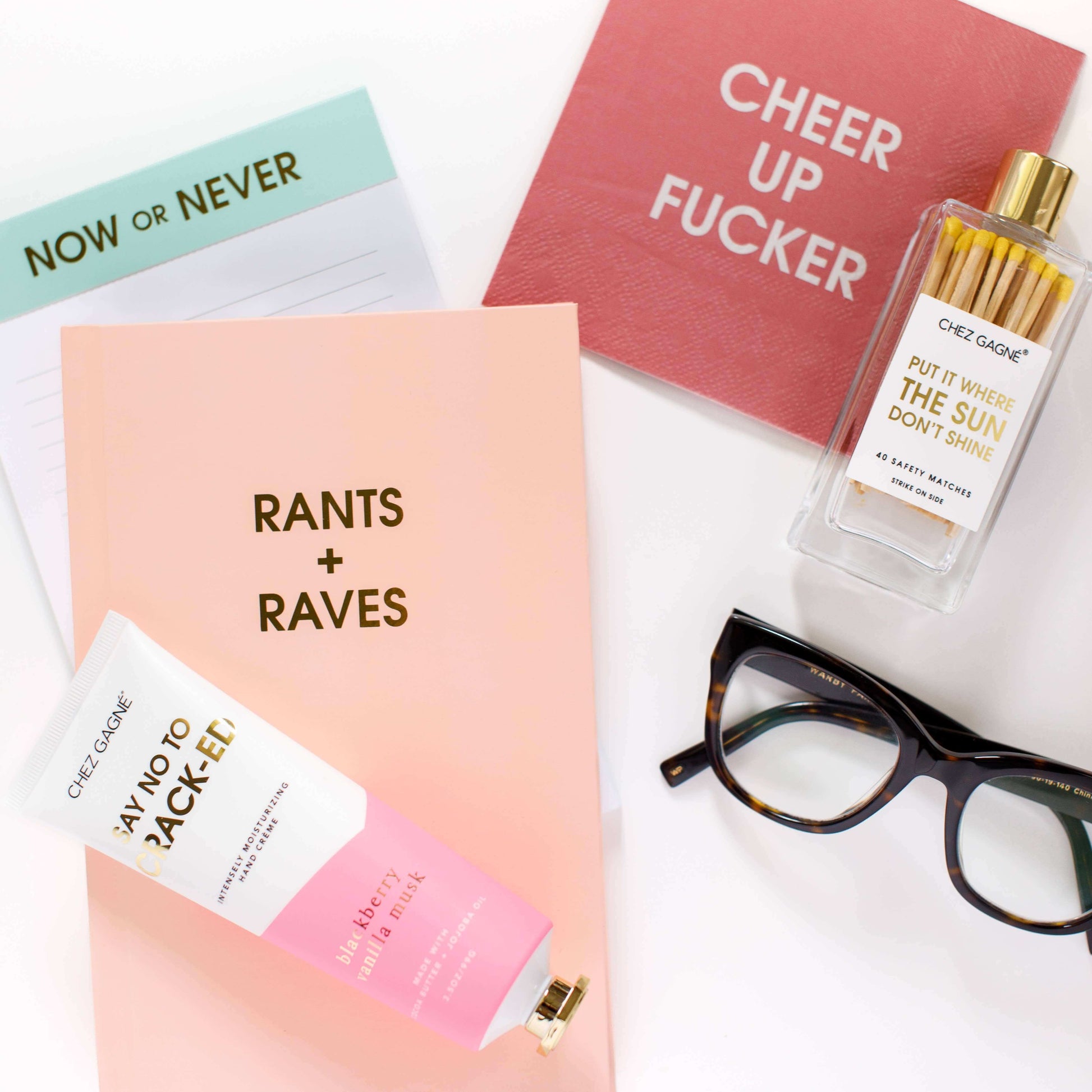 A flat lay of a playful journal collection featuring the Rants + Raves journal in peach, motivational cards, glasses, lotion, and perfume.