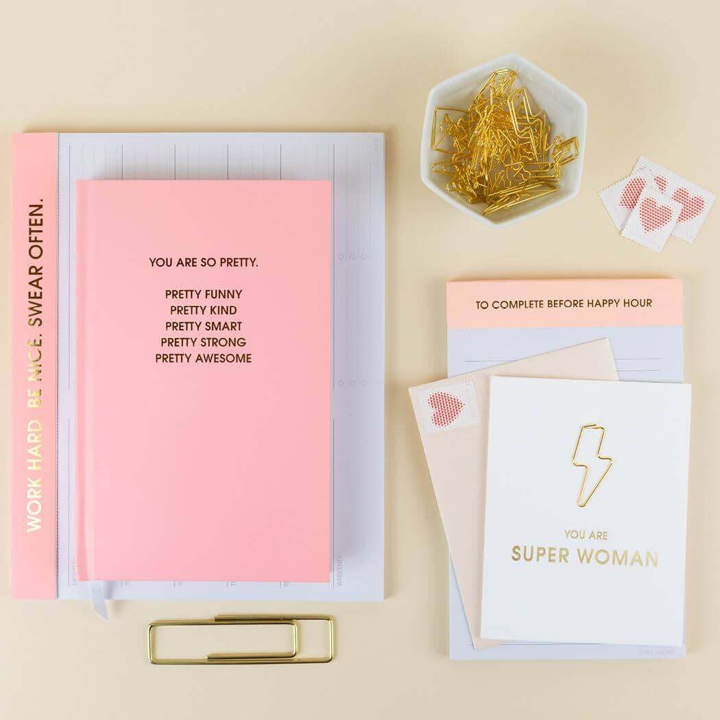 Pretty sunset pink journal with gold foil print, pink cards, golden paper clips, and office supplies on a desk for journaling.