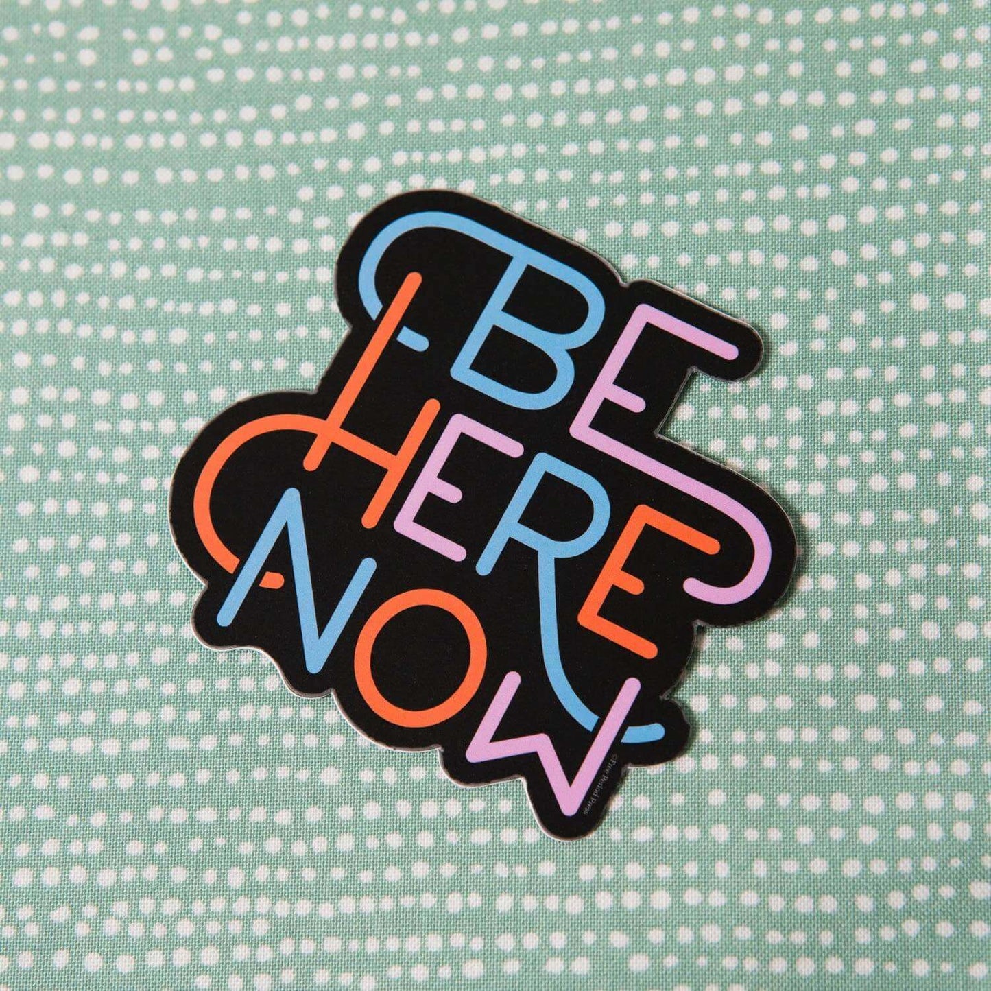 Colorful "Be Here Now" vinyl decal sticker on dotted green background, perfect for journals, laptops, or water bottles.