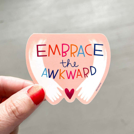 Hand holding "Embrace the Awkward" vinyl decal sticker with colorful text and a heart, perfect for journaling, laptops, and more.