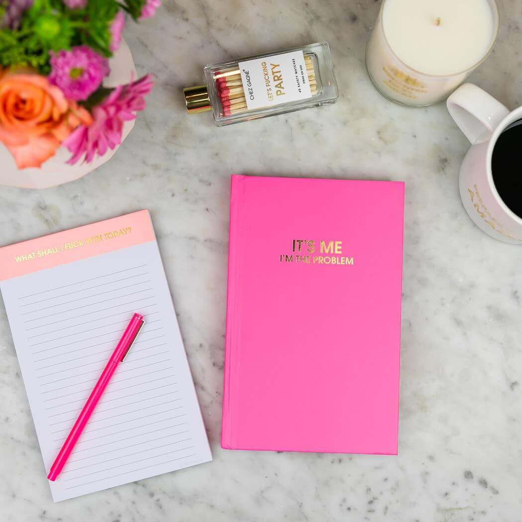 Cosmo Pink "It's Me I'm The Problem" journal with gold foil print on a marble table next to notepad, flowers, and a candle.