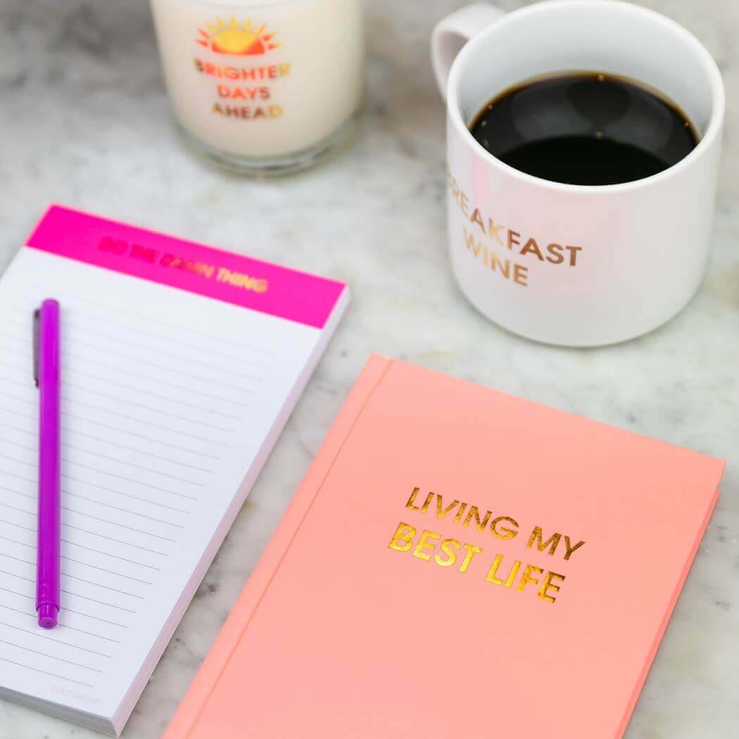 Bellini Pink "Living My Best Life" journal with gold foil, coffee mug, notepad, and candle. Perfect for uplifting journaling sessions.