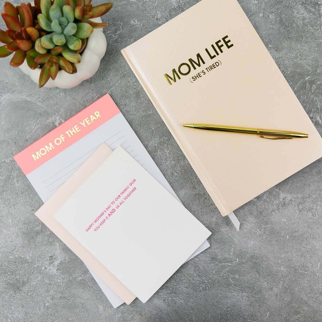 Coral sparkle "Mom Life (She's Tired)" journal with gold foil, pen, and journaling supplies on grey surface with succulent plant.