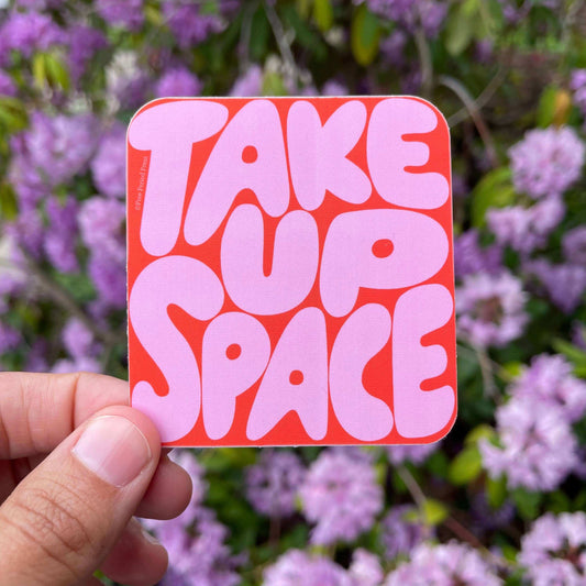 Person holding Take Up Space Vinyl Decal Sticker with bold pink letters against a red background, flowers in the background. Decor for journaling.