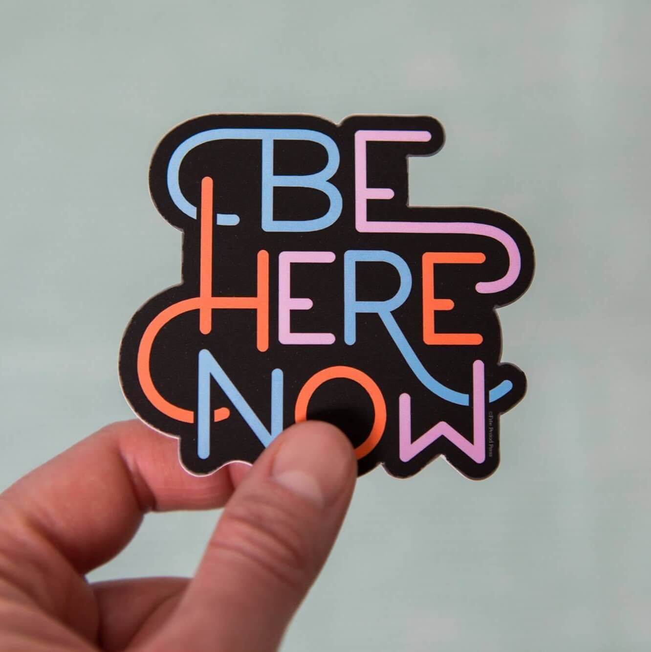 Hand holding colorful "Be Here Now" vinyl decal sticker for journaling, Macbook, planners, skateboard, and Hydroflask – designed by Katie Daugherty.