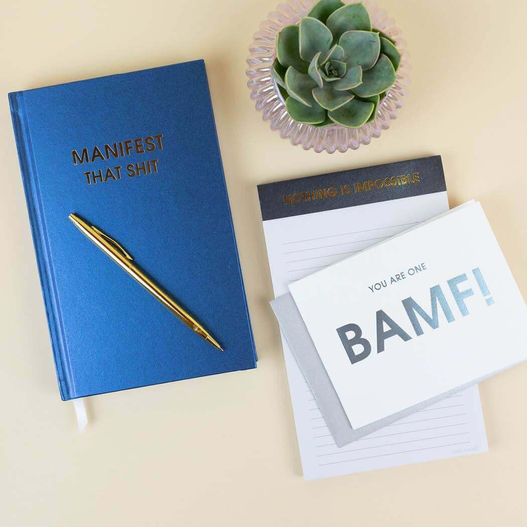 Bright hardcover "Manifest That Shit" journal with gold pen, succulent plant, notebook, and motivational cards for guided journaling.