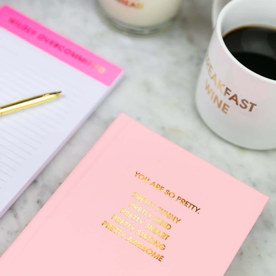 Sunset pink hardcover "You Are So Pretty" journal with gold foil print beside a coffee mug, a notepad, and a pen on marble surface