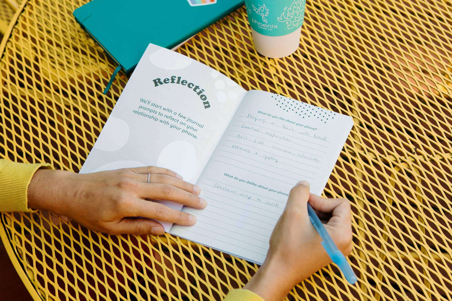 Person journaling in the Unplugged workbook with a reflection page, blue pen, and coffee on a yellow table. Digital detox and reflection journal.