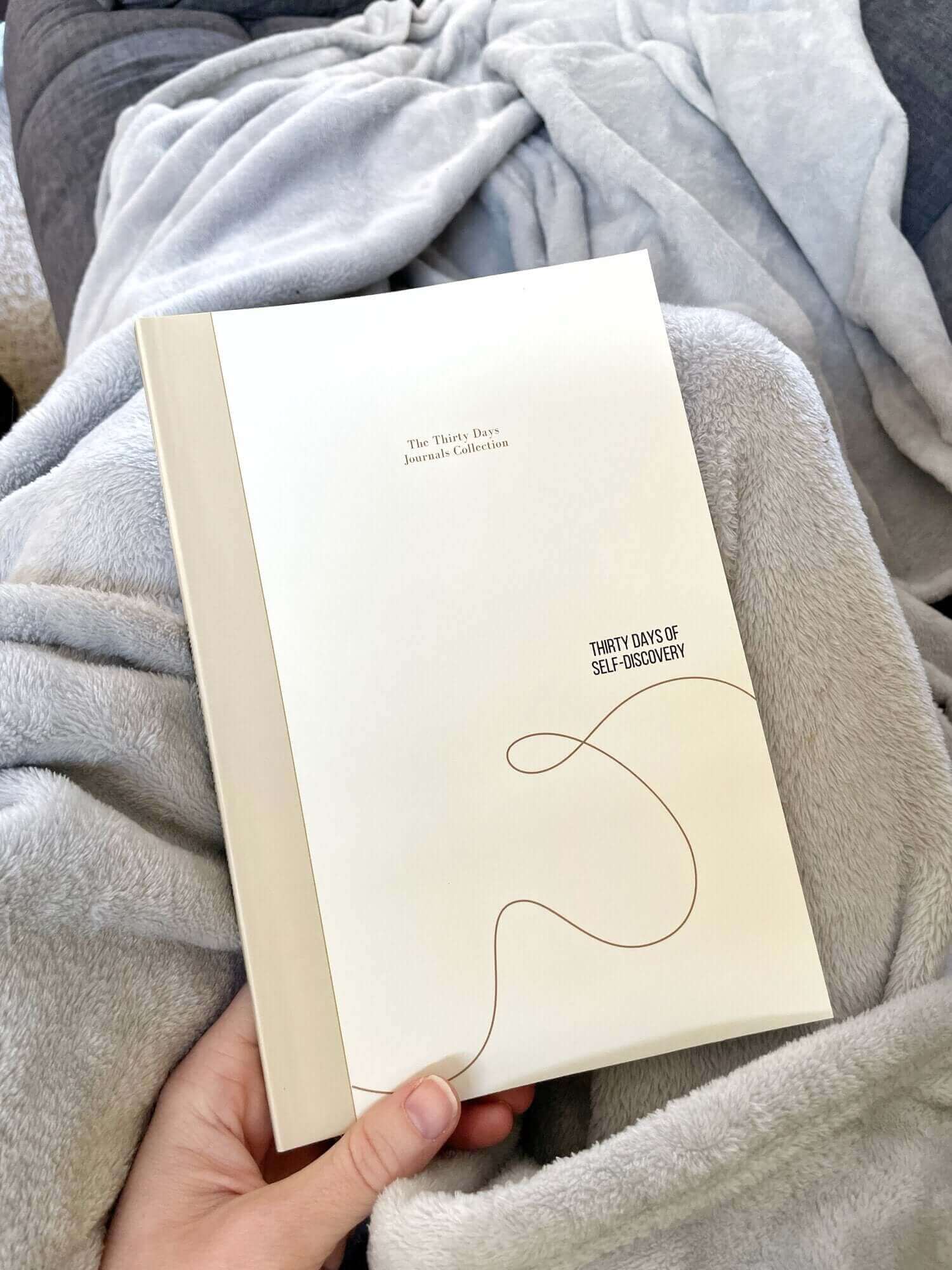 Person holding "Thirty Days of Self-Discovery" journal, wrapped in a cozy blanket, ready for a transformational journaling journey.