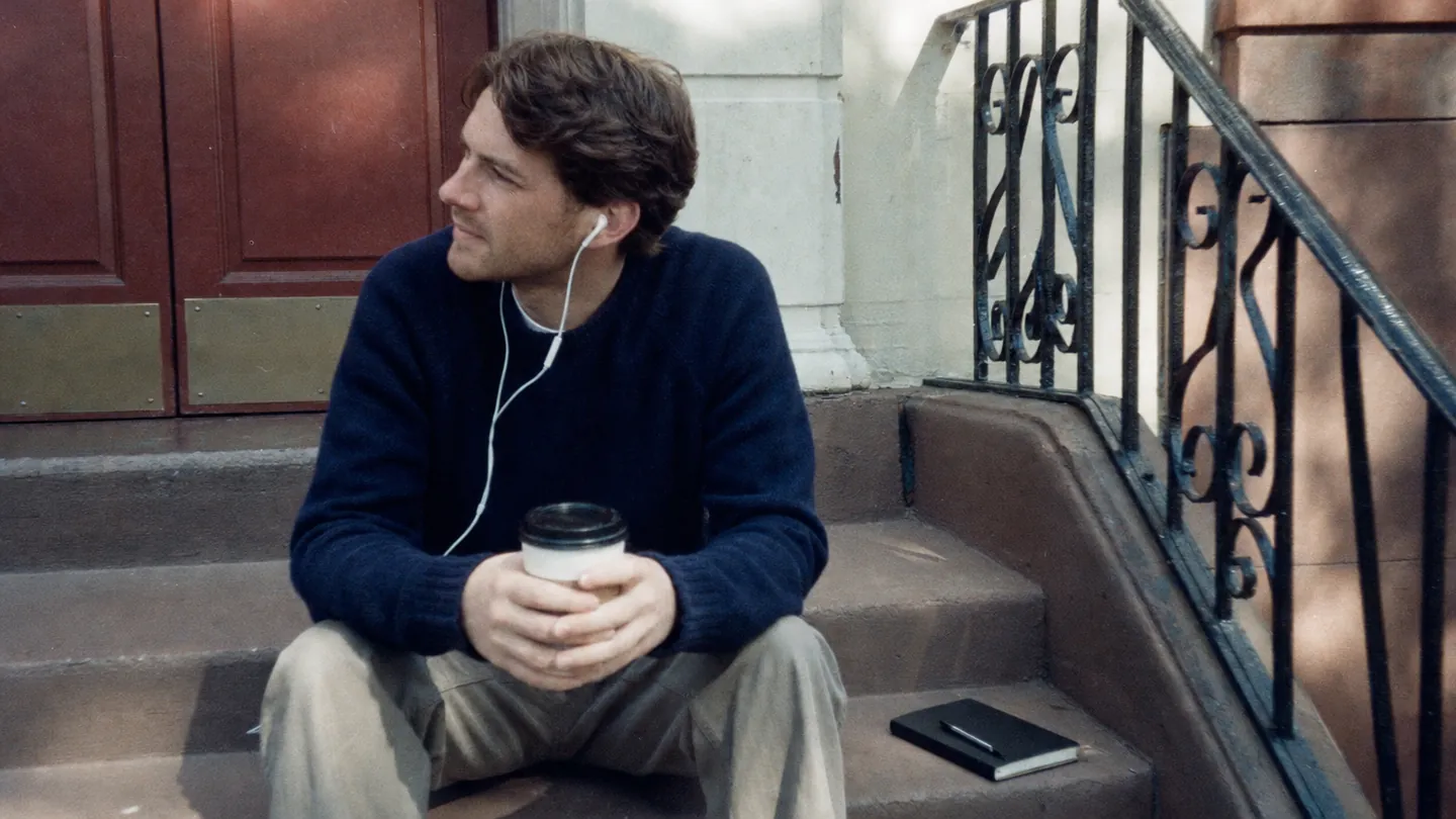 A man sits on his front steps with a coffee and earbuds in, a closed notebook and pen beside him.