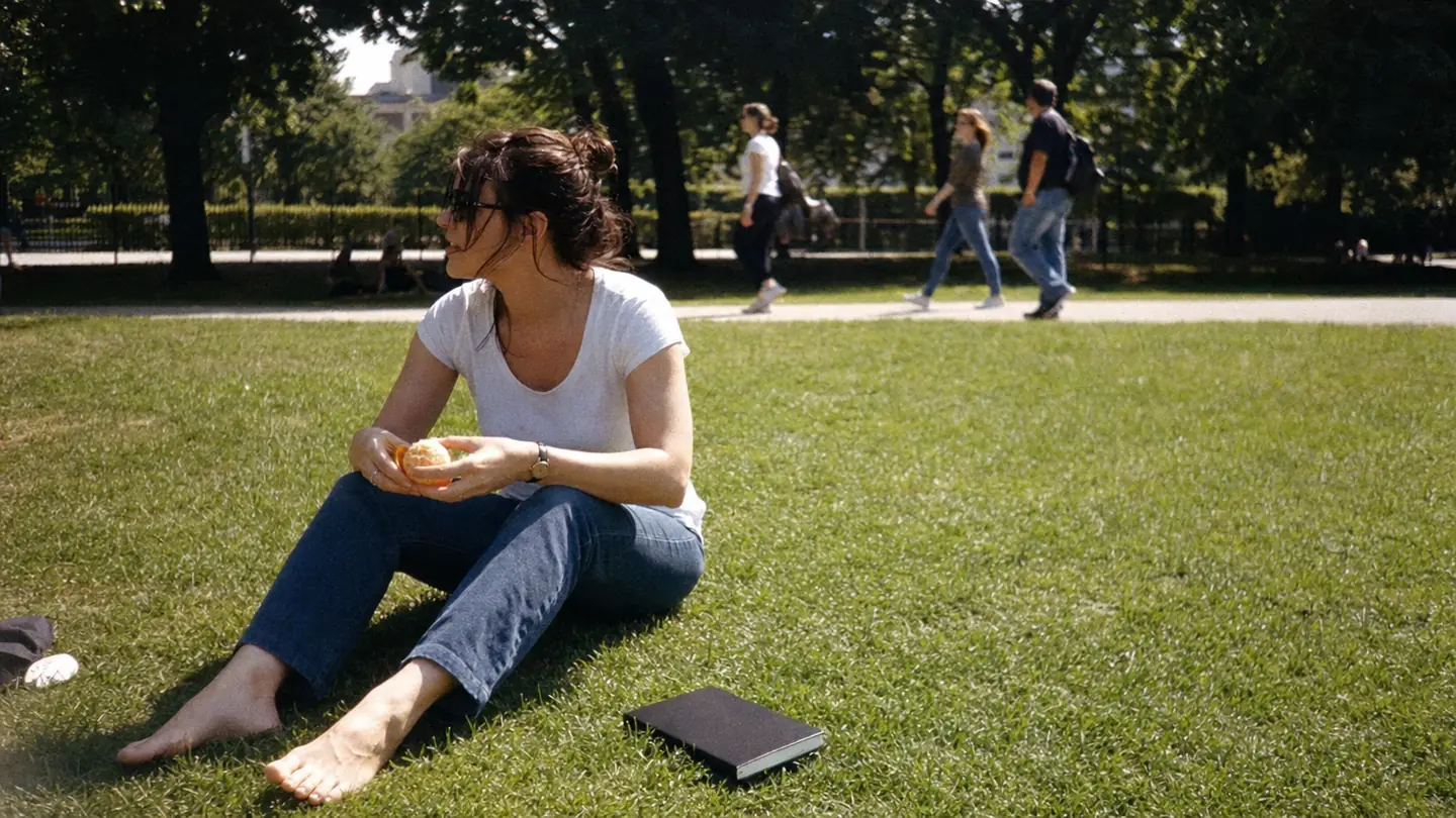 A woman sits barefoot on a park lawn at lunchtime, peeling an orange, a closed notebook on the grass beside her.