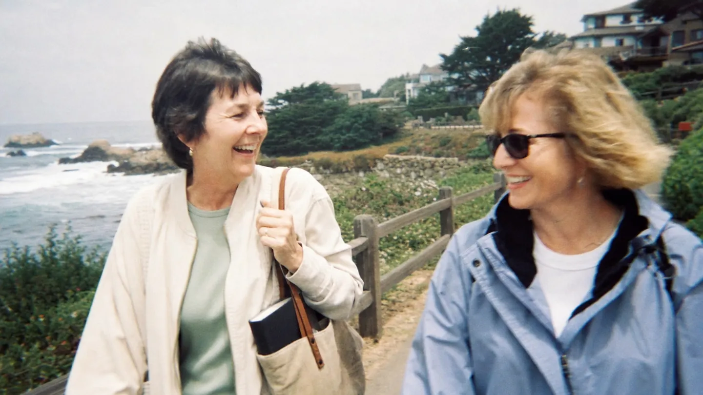 Two women laugh together on a seaside path, a closed notebook in one tote bag.