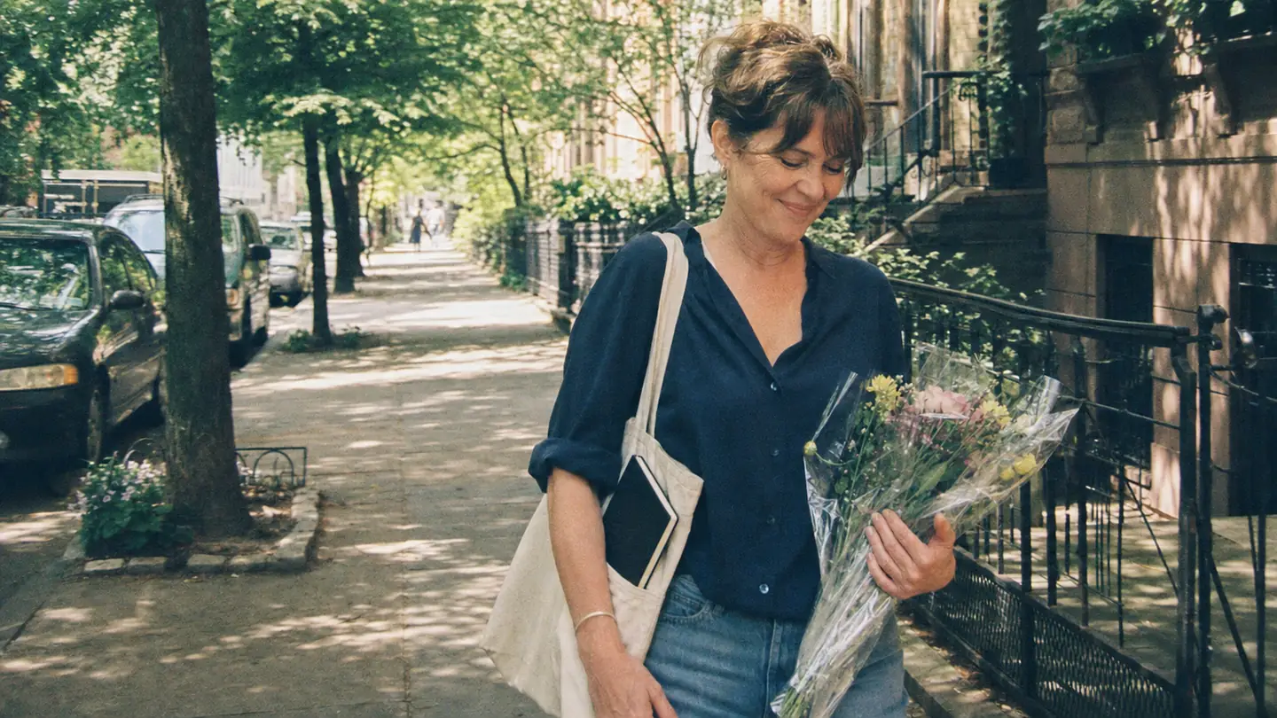A woman walks down a tree-lined street with flowers she bought for herself, a notebook in her tote bag.