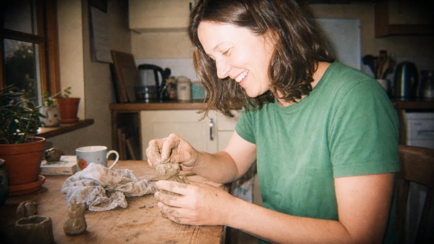 A woman makes a small clay animal at her kitchen table.